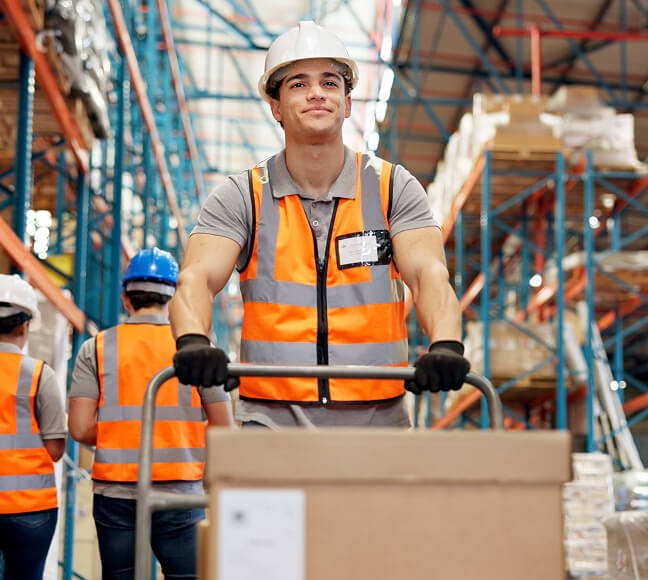 Warehouse worker in an orange safety vest and hard hat pushing a cart loaded with a box through a wholesale warehouse.