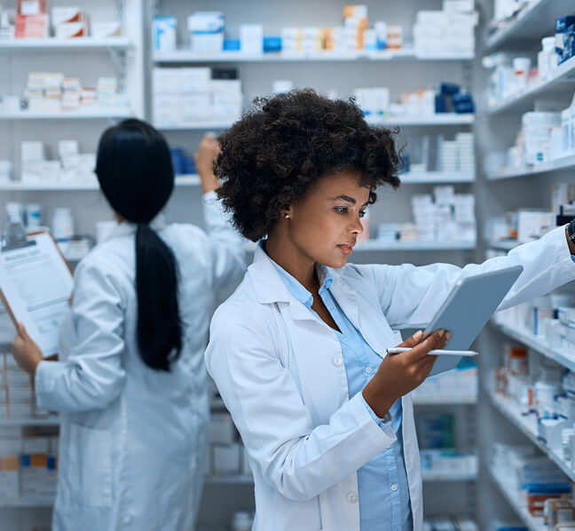 Pharmacist reviewing a patient’s prescription order on a tablet while searching pharmacy shelves for the medication.