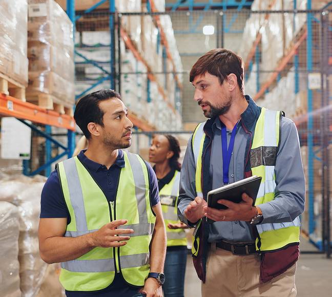 Two manufacturing workers wearing yellow safety vests walking through a warehouse with a clipboard.