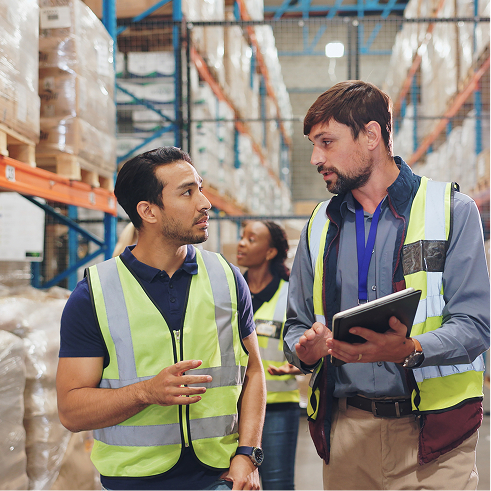 Two manufacturing workers wearing yellow safety vests walking through a warehouse with a clipboard.